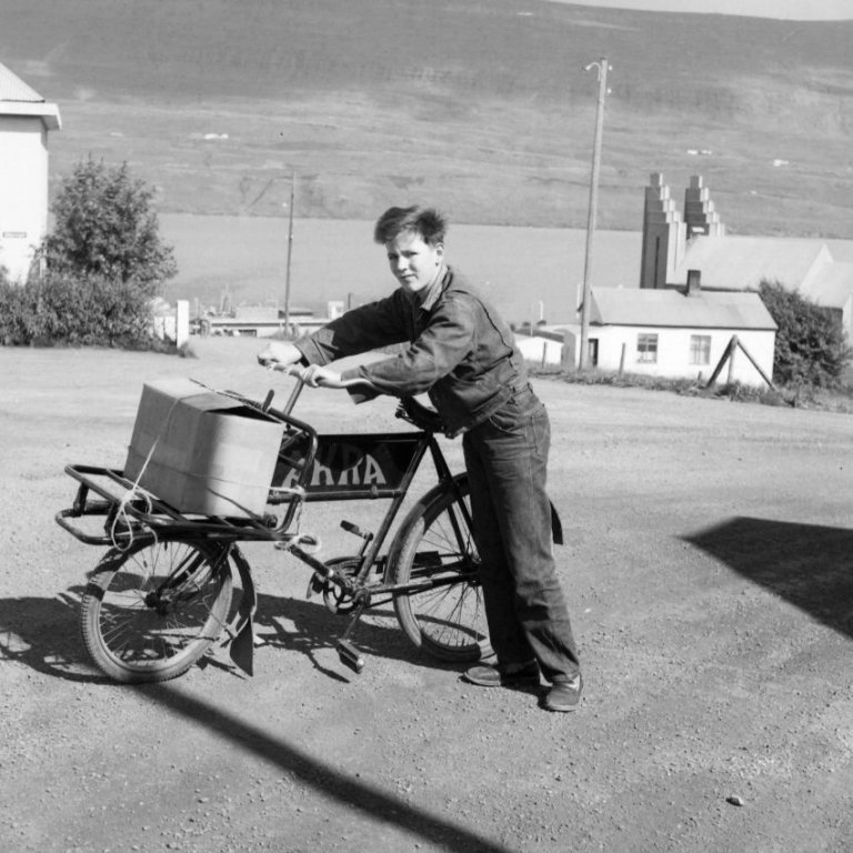1. Sendill frá Smjölíkisgerðinni Akra, um 1960. Mörg fyrirtæki buðu upp á þá þjónustu að senda varning heim til fólks. Ungir hraustir piltar þeyttumst um bæinn með margvíslegan varning framan á bögglaberanum sem var framan á hjólinu. Bak við þennan sendil sést í húsið Garð sem stóð í brekkunni neðan við Frímúrarahúsið. Það var rifið 1967 og hafði þá verið í eigu Akureyrar frá 1955. Ljósmynd Erlingur Davíðsson.