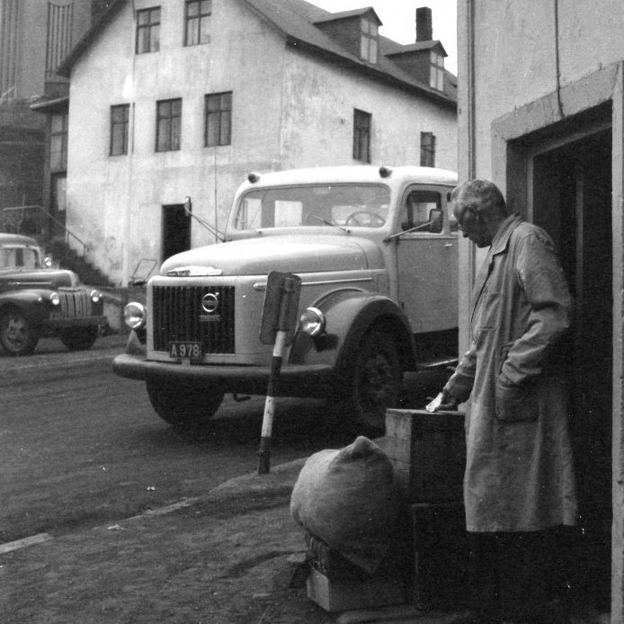 104. Beðið við Bögglageymsluna í Kaupvangsgili, um 1964. Mjólkurbílinn A- ? er handað við hornið. Ef til till hefur bílstjórinn brugðið sér inn í bögglageymsluna til að taka kassa með matvörum út í nærliggjandi sveitir. Algengt var að viðskiptavinir KEA hringdu inn í matvörudeild KEA og pöntuðu vörur sem þeir fengu svo heimsendar með mjólkurbílnum eða jafnvel Dalvíkurrútunni. Hvíta stjóra húsið er gistiheimið Caroline Rest sem amerikaninn George Schrader byggði 1913-1914. Á þessu sérstæða gistiheimili stóð líka hestum til boða gisting en í hesthúshlutanum var hægt að hýsa 130 hesta við stall. Húsið var rifið 1979. Ljósmynd Erlingur Davíðsson.