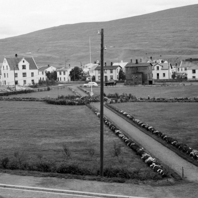 24. Eiðsvöllur á Oddeyri. Eiðsvöllurinn var upphaflega settur inn á Aðalskipulag Akureyrar árið 1927 og átti að vera myndarlegur bæjarvöllur og miðpunktur fyrir íbúa á hinni flötu Oddeyri. Nafnið fékk hann líklega 1928 en lítið varð úr framkvæmdum við völlinn og hann var áfram tún að stærstum hluta. Í seinni heimstyrjöldinni var herbraggakampur inn Valhalla á Eiðsvelli og á myndinni má sjá einn þeirra lengst til vinstri á myndinni. Það var ekki fyrr en vorið 1950 sem gerð vallarins hófst, sáð var grasfræi, lagðir gangstígar og plantað blómum. Árið 1953 stóð Fegrunarfélag Akureyrar fyrir því að reisa hvítu fánastöngina í tilefni af afmæli bæjarins árið áður en þá var ritað í bæjarblöðin að Eiðsvöllur væri einn af fegurstu reitum í bænum. Dökka húsið á vellinum er Norðurgata 27 sem var byggt 1905 en var að lokum rifið. Ljósmynd Gísli Ólafsson.