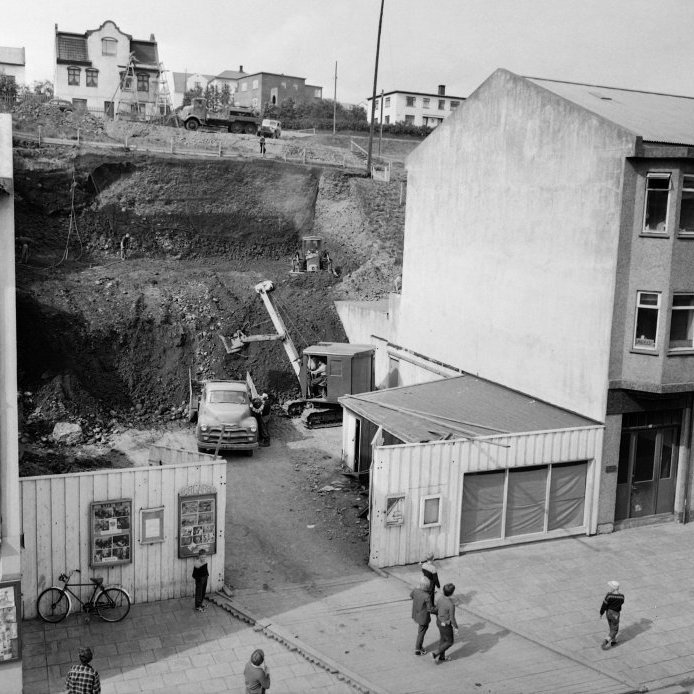26. Stórverslun Amaro í byggingu árið 1959. Grjóthörð móhellan var losuð með lofthömrum áður en víragrafa flutti lausan jarðveginn á vörubílspallinn. Ungur drengur fylgist spenntur með ofan af brúninni. Ljósmynd Eðvarð Sigurgeirsson.