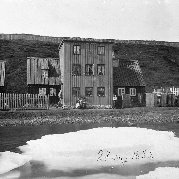 53. Hafnarstræti 23, 28. ágúst 1882. Húsið byggði ein merkasta og umtalaðasta kona sinnar samtíðar á Akureyri, Vilhelmina Lever. Hún rak verslun í húsinu en síðar keypti Höepfner kaupmaður það og stofnaði þar fyrsta bakaríið í bænum árið 1867. Anna Schiöth tók myndina af húsinu og maður hennar, Hendrik Schiöth bakari stendur við húsið og börnin þeirra sitja fyrir framan. Myndin er tekin í lok ágúst og þetta ár var hafís í Eyjafirði allt sumarið sem olli hungursneyð hér. Húsið brann 1903. Ljósmynd  Anna Schiöth.