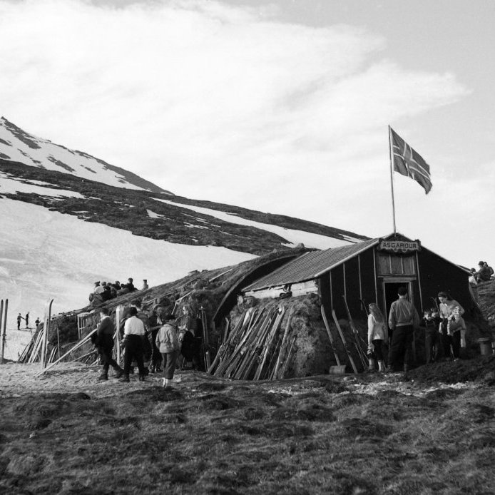 72. Skíðaskálinn Ásgarður, um 1950, sem stóð nokkru sunnan við Skíðahótelið. Þennan skála reistu nemendur Gagnfræðaskólans og Iðnskólans en árið 1944 keyptu þeir í sameiningu gamlan hermannaskála og fluttu hann á upp í fjall og reistu hann aftur undir stjórn Hermanns Stefánssonar leikfimikennara. Byggt var inngangur framan á braggann og torfveggir voru hlaðnir meðfram hliðum beggja til húsanna til einangrunar. Það kom fyrir að bragginn færi nánast á kaf í snjó. Þessi skíðaskáli var aflagður þegar Skíðahótelið var tekið í notkun.  Ljósmynd Kristján Hallgrímsson.