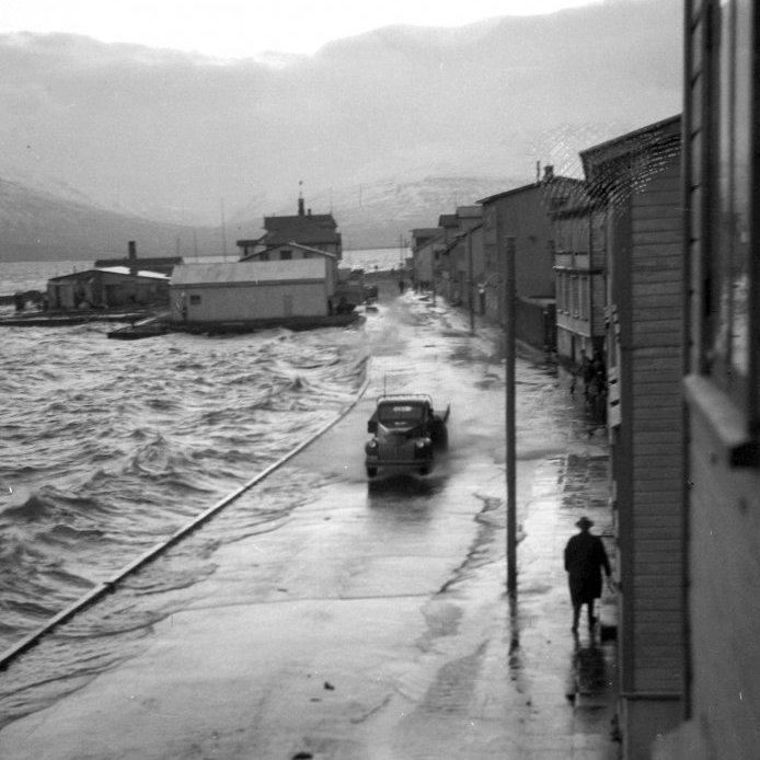 82. Hafnarstrætishúsin um 1950. Myndin er tekin af svölunum á Hafnarstræti 41. Í stórstraumsflóði og stífri austanátt gekk sjórinn yfir götuna og flæddi inn í kjallara húsanna. Ljósa húsið sem snýr langhlið fram er flugskýli Flugfélags Akureyrar sem var byggt árið 1937-38. Svæðið í dag er mikið breytt vegna seinna tíma landfyllinga. Ljósmynd Kristján Hallgrímsson.