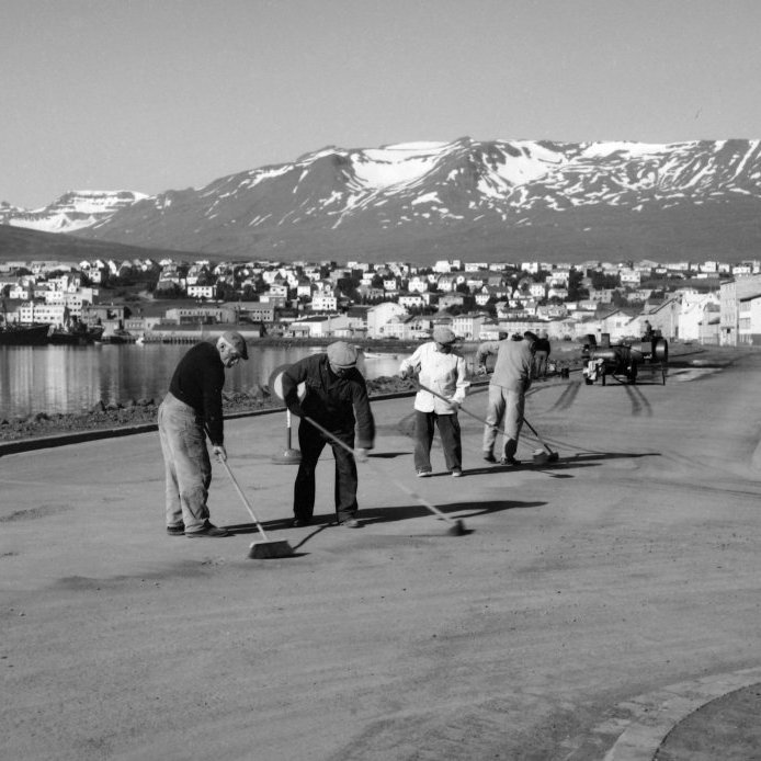 83. Vinnuflokkur bæjarstarfsmanna við malbiksviðgerðir á Strandgötu um 1950. Þegar fyrstu götur voru lagðar malbiki var það lagt út með hrífum. Bikið var því ójafnt og misheitt þannig að frostskemmdir voru algengar, jafnvel á nýju malbiki. Ljósmynd Eðvarð Sigurgeirsson.
