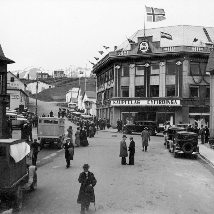 98. Kaupvangstræti árið 1936. Tilefnið er 50 ára afmæli KEA og er stórhýsi KEA frá 1930 er skreytt í tilefni dagsins. Leigubílaröðin bíður eftir því að ferja gesti í afmælissamsæti KEA sem haldið var á Hrafnagili. Ljósmynd Vigfús Sigurgeirsson.