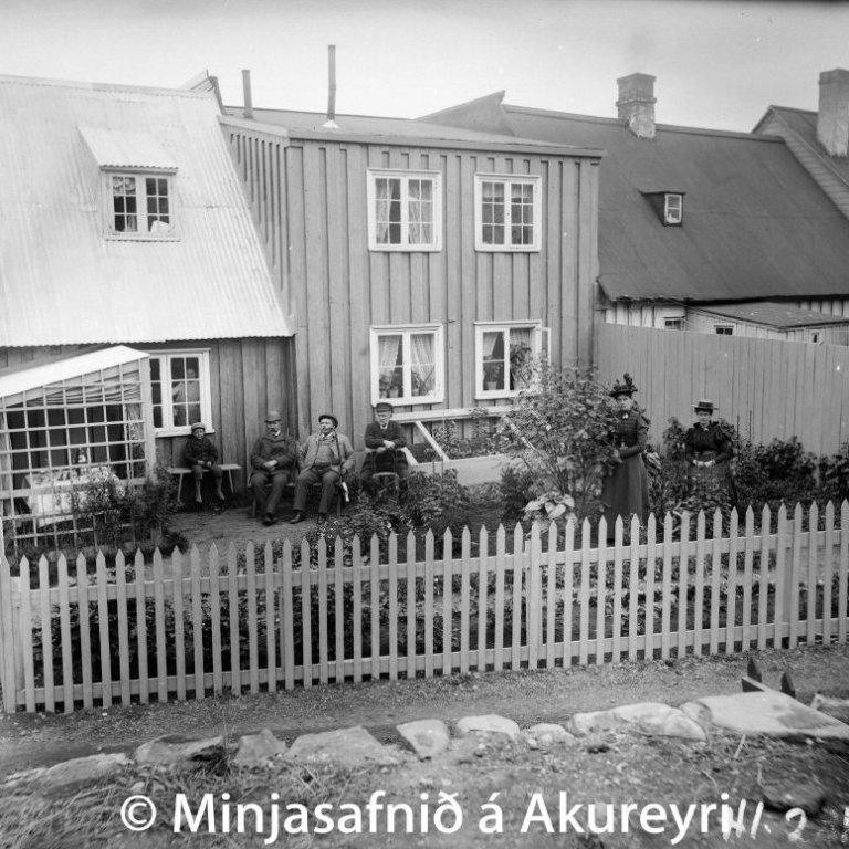 Garður Hendriks Schiöth, póstmeistara, og Olgu Schiöth konu hans við Aðalstræti 6.
Á myndinni situr ungur drengur á trébekk. Þar við hliðina er óþekktur maður en við hlið honum situr Oddur C. Thorarensen, þá Hendrik Schiöth, Alma Thorarensen og Olga Schiöth. Búðarlækurinn rennur þarna framhjá og sést grjóthleðslan á vesturbakkanum.