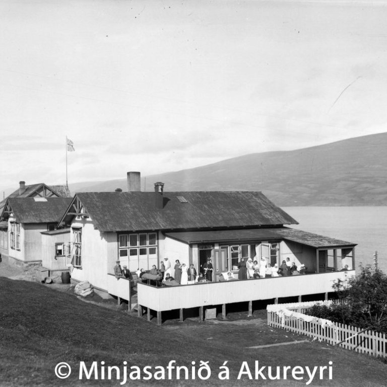 Spítalinn séður frá suðvestri. Sjúklingar og starfsfólk nýtur sólar á pallinum spítalans. Stóri glugginn, hvítur að neðan, var á skurðstofunni. Sunnan við trjágarðinn var síðar byggt sérstakt sólbaðsskýli fyrir sjúklinga.